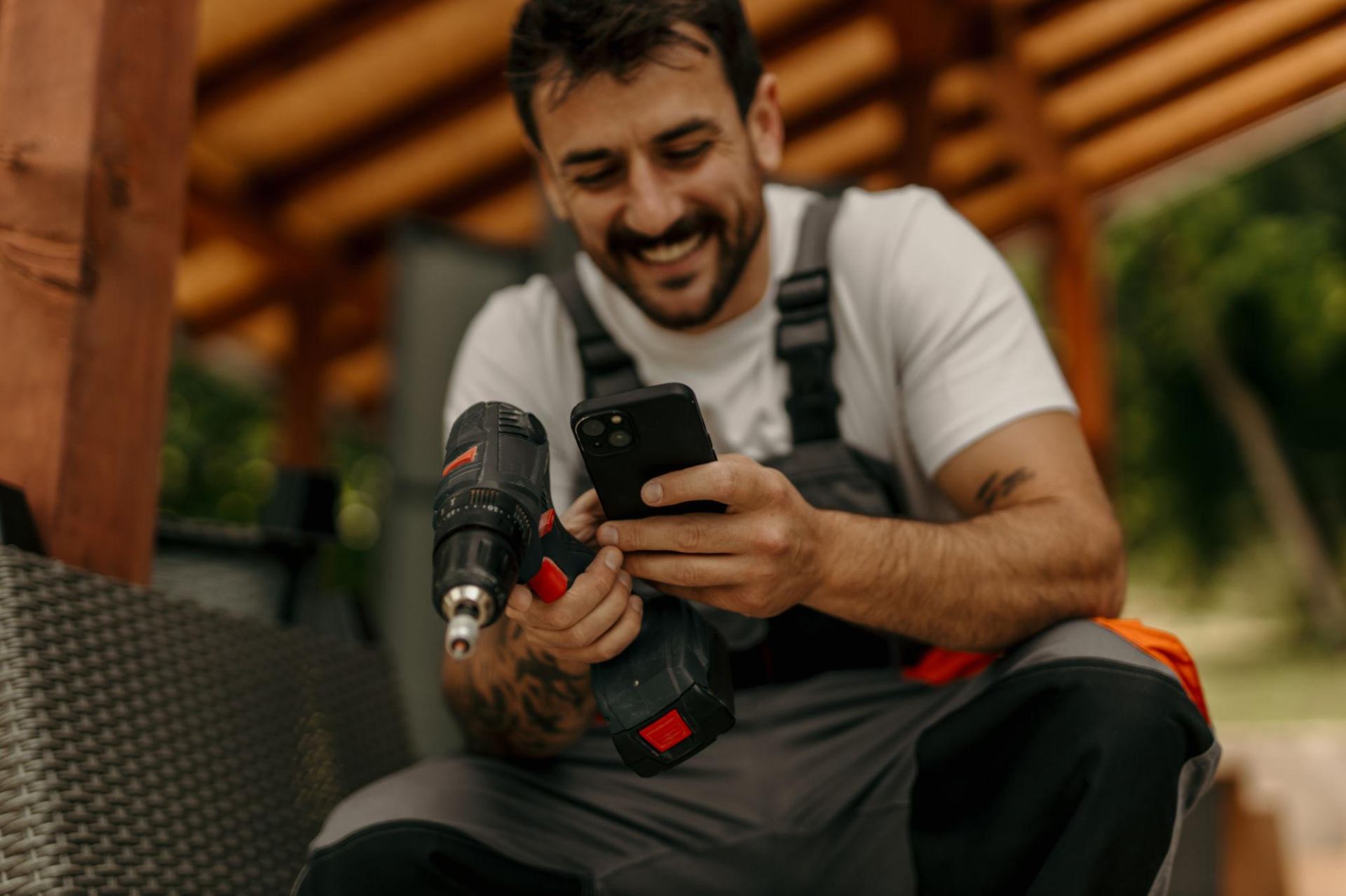 Smiling handyman in work overalls holding a cordless drill and checking his phone on a job site